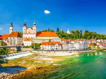 Blick auf Steyr, Oberösterreich, mit barocken Kirchtürmen, bunten historischen Gebäuden und einem Flusswehr unter blauem Himmel.