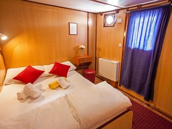 Gemütliche Kabine auf der MS Azimut mit Doppelbett, roten Kissen, holzgetäfelten Wänden, Bullauge mit blauen Vorhängen und weißer Heizung.