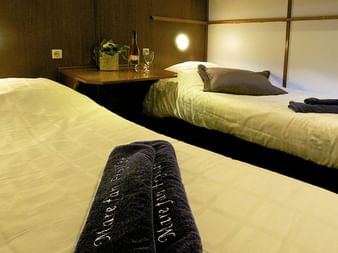 Zweibettkabine auf Mare fan Fryslân mit dunkler Holzvertäfelung, Wandleuchten und Nachttisch mit Weinflasche und Gläsern zwischen den Betten.