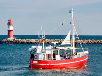 Rot-weißes Passagierboot fährt nahe einem rot-weiß gestreiften Leuchtturm im Rostocker Hafen. Steinmole und blaues Meer sichtbar.