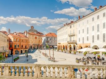 Piazza Maggiore in Mondovì mit historischen Gebäuden, Arkadenfassaden, Straßencafés und steinerner Balustrade im Vordergrund unter blauem Himmel.