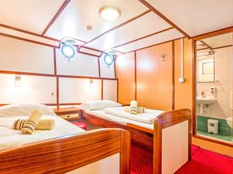 Doppelkabine auf dem Schiff Amore mit zwei Einzelbetten, Holzmöbeln, Bullaugen, rotem Teppich und eigenem Bad mit Waschbecken.