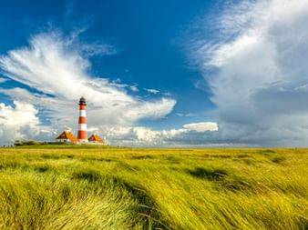 Leuchtturm Westerhever mit rot-weißen Streifen und Nebengebäuden in grüner Salzwiese unter dramatischem Wolkenhimmel an der Nordsee.