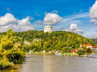 Befreiungshalle auf bewaldetem Hügel über Kelheim mit Donau, Dorfkirche und Ausflugsschiffen unter blauem Himmel mit Wolken.