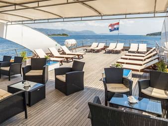 Geräumiges Sonnendeck der MS Olmissum mit Holzboden, Sonnenliegen, Korbmöbeln und kroatischer Flagge. Küstengebirge im Hintergrund sichtbar.