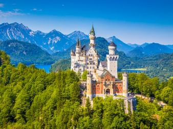 Schloss Neuschwanstein umgeben von grünen Wäldern mit Alpenbergen und blauen Seen im Hintergrund unter klarem blauen Himmel bei Füssen.