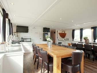 Geräumiger Salon an Bord der MS Magnifique II mit großem Holzesstisch, braunen Lederstühlen und weißem Küchenbereich. Große Fenster mit Vorhängen an beiden Seiten.
