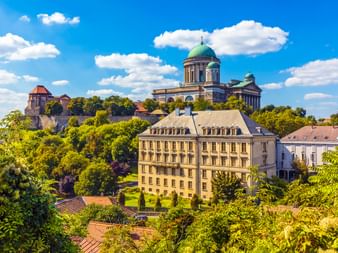 Esztergom Basilika mit grüner Kuppel auf einem Hügel über historischen Gebäuden und üppiger Vegetation unter blauem Himmel mit weißen Wolken.