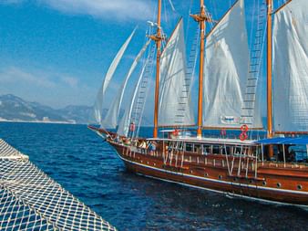 Holzsegelschiff MS Thalassa mit weißen Segeln auf blauem Meer, von einem anderen Schiff aus gesehen. Berge im Hintergrund unter klarem Himmel.