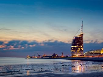 Abendansicht des Bremerhavener Hafens mit beleuchtetem segelförmigem Gebäude und goldkuppelförmigem Klimahaus, das sich im nassen Strandsand spiegelt.
