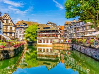 Traditionelle Fachwerkhäuser spiegeln sich in einem Kanal in Straßburg. Bunte Blumen schmücken die Steinbrückengeländer unter blauem Himmel.