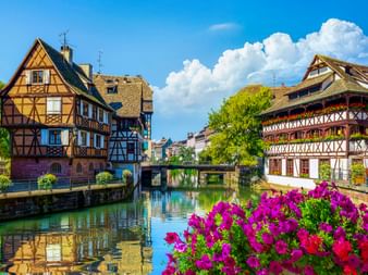 Traditionelle Fachwerkhäuser entlang eines Kanals in Petite France, Straßburg, mit bunten Blumen im Vordergrund und blauem Himmel mit Wolken.