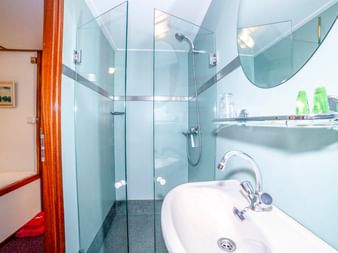 Kompaktes Badezimmer auf der MS Lena Maria mit weißem Waschbecken, verglaster Dusche, hellblauen Fliesen und Toilettenartikeln im Regal.