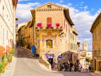 Steingebäude in Assisi mit religiösem Fresko und Balkon mit rosa Blumen. Enge Straße führt aufwärts mit Geschäften unten und Glockenturm.