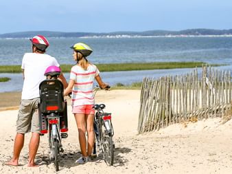 Drei Kinder mit Fahrrädern und Helmen auf sandigem Strandweg an der Ostsee, Blick auf Küstenlandschaft mit Wasser und ferner Küste.