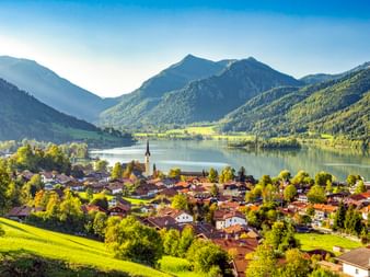 Luftaufnahme des Dorfes Schliersee in Bayern mit Kirchturm, umgeben von See und bewaldeten Bergen unter blauem Himmel.