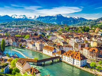 Luftaufnahme der Luzerner Altstadt mit bunten Gebäuden entlang der türkisfarbenen Reuss, Brücken über das Wasser und schneebedeckten Alpen.