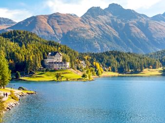 Historisches Waldhaus Hotel auf einer Halbinsel in einem blauen Bergsee bei St. Moritz, umgeben von Wäldern und Alpengipfeln unter klarem Himmel.