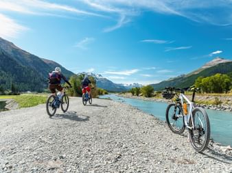 Two cyclists riding on a gravel path beside the turquoise Inn River in Austria, with mountains and forests in the background under blue sky.