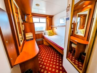 Kompakte Einzelkabine auf MS Rigoletto mit Einzelbett, Fenster mit Meerblick, Holzmöbeln, rot gemustertem Teppich und Spiegel.