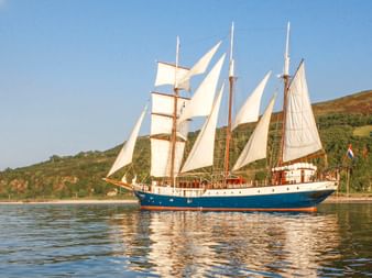 Dreimastiger Großsegler Atlantis mit weißen Segeln, segelnd auf ruhigem Wasser mit grünen Hügeln im Hintergrund unter blauem Himmel.