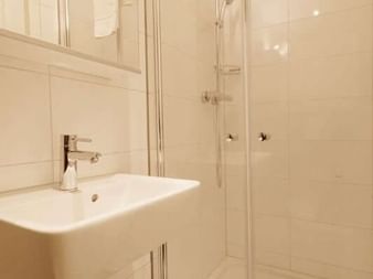 Kompaktes Badezimmer auf der MS Magnifique III mit weißem Waschbecken, Spiegel, Glasduschkabine und beigen Fliesen.