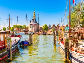 Traditionelle Boote im Hafen von Hoorn mit historischem Turm und Giebelhäusern unter blauem Himmel. Holzpfähle säumen die Wasserstraße.