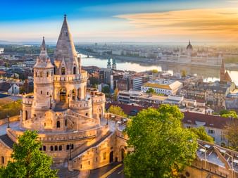 Türme der Fischerbastei in Budapest bei Sonnenuntergang mit Donau und ungarischem Parlamentsgebäude im Hintergrund.