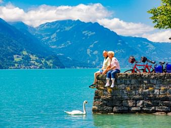Zwei Radfahrer sitzen auf einer Steinmauer am Brienzersee, ein weißer Schwan schwimmt in der Nähe. Berge und blauer Himmel im Hintergrund.