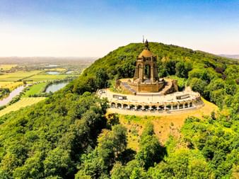 Luftaufnahme des Kaiser-Wilhelm-Denkmals auf einem bewaldeten Hügel in Porta Westfalica, mit der Weser, die durch grüne Felder fließt.