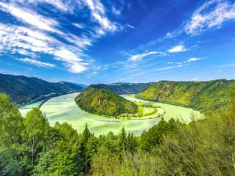 Luftaufnahme der Schlögener Schlinge, einer Flussschleife der Donau, umgeben von bewaldeten Hügeln unter blauem Himmel mit weißen Wolken.