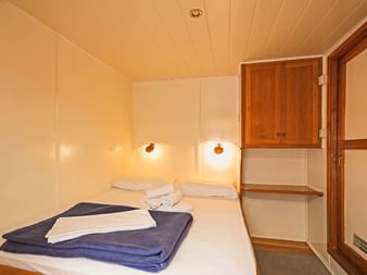 Kompakte Kabine auf der MS Sveti Vid mit weißem Doppelbett, blauen Handtüchern, Wandleuchten, Holzschrank und Tür mit Milchglasscheiben.