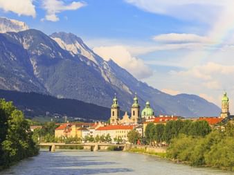 Blick auf den Inn, der durch Innsbruck fließt, mit historischen Kirchtürmen und Gebäuden. Dramatische Alpenberge erheben sich im Hintergrund unter blauem Himmel.