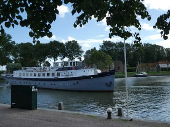 Passagierschiff Liza Marleen mit dunkelblauem Rumpf und weißem Aufbau im Kanal vertäut, umgeben von Bäumen und Gebäuden unter blauem Himmel.