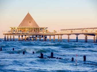 Seebrücke Heringsdorf auf Usedom mit markanter pyramidenförmiger Überdachung, die sich in die Ostsee erstreckt. Wellen umspülen Holzpfosten.