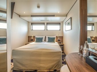 Moderne Kabine auf MS Swiss Crown mit Doppelbett, beiger Bettwäsche, türkisfarbenen Kissen, Holznachttischen, Wandlampen, Fenstern und Spiegel.
