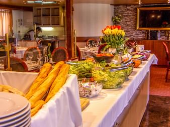 MS Olympia Restaurant mit Buffettisch mit Baguettes, Salaten und Tulpen. Speisebereich mit weißen Tischdecken und Holzstühlen.