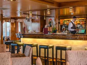 MS Olympia Schiffsbar mit beleuchtetem Holztresen, Barhockern, beigen Sesseln und Barkeeper. Bullaugen und warmes Licht schaffen gemütliche Atmosphäre.