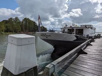 Flusskreuzfahrtschiff Floor van Aemstel (ehemals Liza Marleen) am Holzsteg mit niederländischer Flagge, weißem Aufbau und dunklem Rumpf.