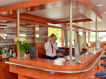 Moderne Schiffsbar auf der MS Fluvius mit Holztheke, Chromsäulen und Barkeeperin im weißen Hemd. Große Fenster zeigen Flussblick.