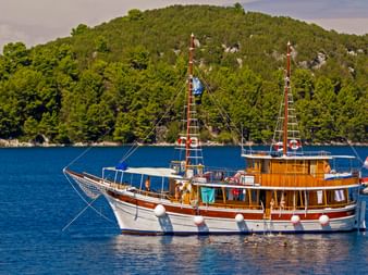MS Orkan, eine traditionelle Motoryacht aus Holz mit weißem Rumpf und Holzaufbau, vor Anker in blauem Wasser mit bewaldeten Hügeln im Hintergrund.