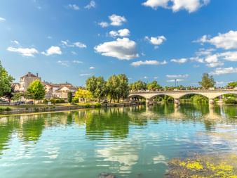 Stone arch bridge spanning the Charente River in Cognac, France. Green water reflects trees and blue sky with white clouds.
