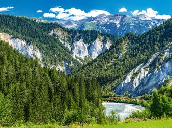 Ruinaulta-Schlucht mit weißen Kalksteinfelsen, dichten grünen Wäldern, einem Fluss im Tal und schneebedeckten Bergen unter blauem Himmel.
