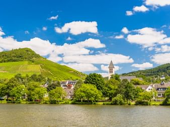 Bullay an der Mosel mit Kirchturm, umgeben von grünen Weinbergen an Hängen unter blauem Himmel mit weißen Wolken.