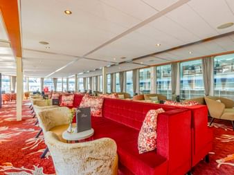 Geräumige Lounge an Bord der MS Lisabelle mit rot gemustertem Teppich, roten und beigen Sofas, großen Fenstern mit Vorhängen und weißer Decke.