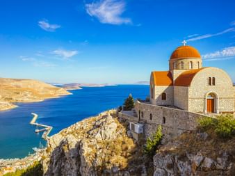 Kloster Agios Savvas mit oranger Kuppel auf felsiger Klippe über dem Hafen Pothia und dem blauen Ägäischen Meer unter klarem Himmel.