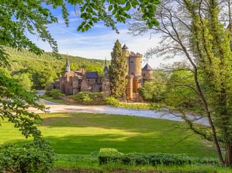 Löwenburg im Bergpark Wilhelmshöhe Kassel, eingerahmt von grünen Bäumen. Das Schloss im mittelalterlichen Stil mit Türmen steht neben einem Teich.