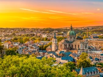 Luftaufnahme der Kathedrale von Namur mit grüner Kuppel und Glockenturm über den Dächern der Stadt unter lebendigem orangefarbenem Sonnenuntergangshimmel.