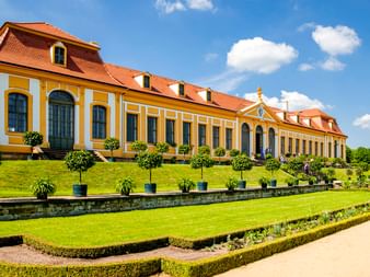 Gelbes Barockschloss mit rotem Dach im Barockgarten Großsedlitz bei Pirna. Gepflegter Rasen mit Zierbäumen und formalen Hecken im Vordergrund.