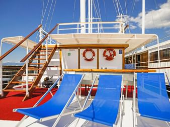 Sonnendeck der MS Sveti Vid mit blauen Liegen auf rotem Boden, weißen Geländern, Holztreppen und orangen Rettungsringen unter blauem Himmel.